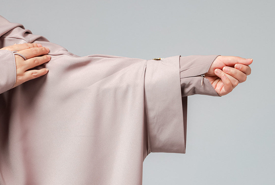 A close-up of a person wearing a flowing, light dusty pink or mauve abaya, showing the fabric texture and sleeve details against a solid gray studio background.